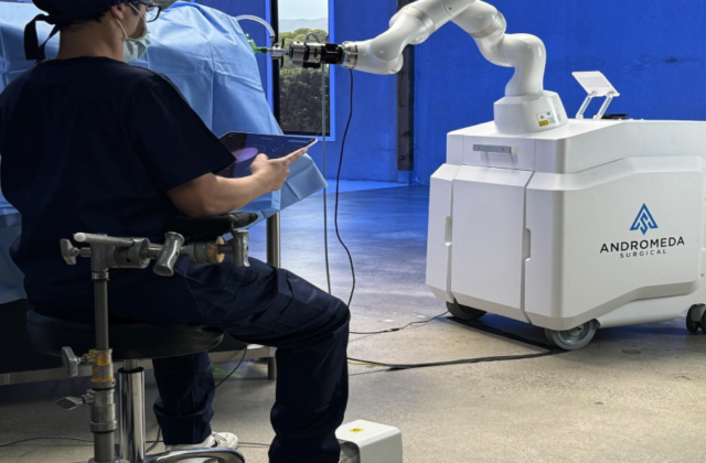 A medical professional operates a robotic surgical system from a tablet during a procedure. The white robotic unit, labeled “Andromeda Surgical,” extends an articulated arm toward the surgical table, demonstrating advanced robotic-assisted surgery technology in a clinical setting.